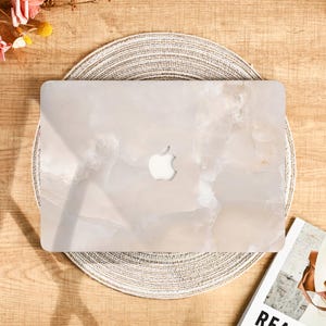 Peut inclure: Un ordinateur portable clair avec un motif semblable &agrave; du marbre, orn&eacute; du logo Apple. L'ordinateur est pos&eacute; sur un dessous-de-plat rond et tiss&eacute;. L'esth&eacute;tique g&eacute;n&eacute;rale est neutre et moderne.