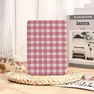 May include: A rectangular case with a pink and white gingham pattern. The case is standing on a woven, circular placemat. The background includes a book with the text "None has begun to think" and a small plant.
