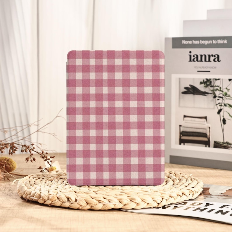 May include: A rectangular case with a pink and white gingham pattern. The case is standing on a woven, circular placemat. The background includes a book with the text "None has begun to think" and a small plant.