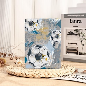 以下が含まれることがあります： サッカーボールのデザインが施された長方形のケース。ケースは、青と黄色の筆致が施されたグレーの背景です。いくつかのサッカーボールが表面に散らばっており、スポーティーなテーマを作り出しています。