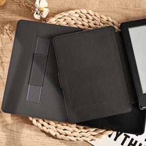 May include: A black e-reader with a matching dark gray case, open to display the screen. The case has a built-in stand and a secure strap. The e-reader is placed on a woven mat, suggesting a focus on reading and technology.
