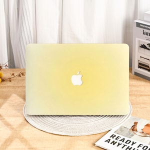 May include: A pale yellow laptop computer with the Apple logo on the lid. The laptop is on a round, woven placemat. The background includes a white curtain and a book.