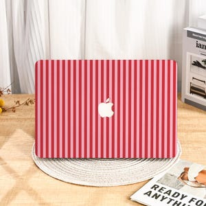 Puede incluir: Funda para portátil con un patrón de rayas verticales en tonos rojos y rosas. El logotipo de Apple está centrado en la funda. La funda está sobre un salvamanteles redondo y tejido.