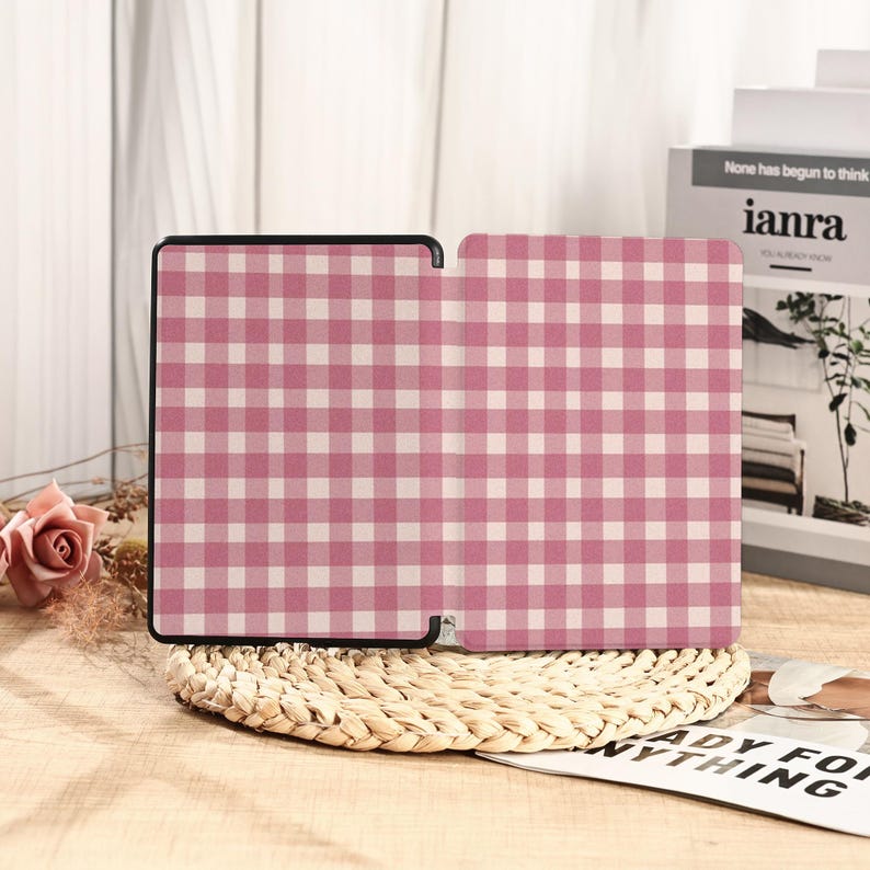 May include: A pink and white gingham pattern tablet case, open to display the interior. The case is on a woven coaster, with a book and a rose in the background. The case has a black border.