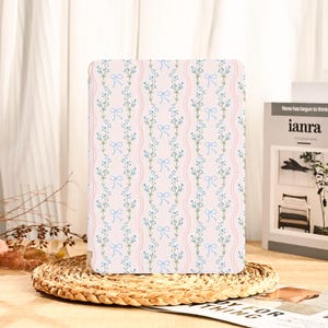 May include: A light pink tablet case with a floral pattern of blue flowers and bows. The case is resting on a woven coaster. The background includes a white curtain and a book with the text "ianra".