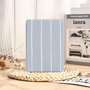 May include: A light blue tablet case with vertical white stripes. The case is rectangular and has rounded corners. It is resting on a woven placemat. In the background, a book with the text "ianra" is visible.