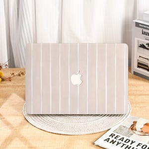May include: A beige laptop case with a white vertical stripe pattern. The Apple logo is visible in the centre. The case is on a round, woven placemat.
