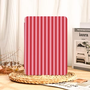 Könnte beinhalten: Eine Tablet-Hülle mit vertikalen Streifen in abwechselnden Rosa- und Rottönen. Die rechteckige Hülle besteht aus glattem Material. Sie liegt auf einem runden, gewebten Platzset. Ein stilvolles Accessoire.