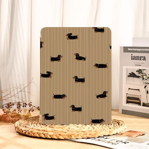 Könnte beinhalten: Eine Tablet-Hülle mit einem sich wiederholenden Muster aus schwarzen Dackel-Hunden auf einem beigefarbenen Hintergrund mit vertikalen Streifen. Die Dackel sind in einem einfachen Cartoon-Stil dargestellt. Die Hülle wird auf einer gewebten Matte präsentiert.