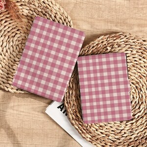 May include: Two notebooks with a pink and white gingham pattern are displayed on a woven surface. The notebooks are open, revealing the pattern on the cover. The background includes a natural woven texture.