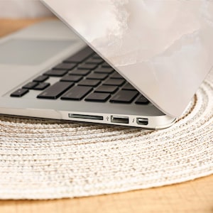 Peut inclure: Un ordinateur portable argent&eacute; avec un clavier noir, partiellement ouvert, r&eacute;v&eacute;lant l'&eacute;cran. L'ordinateur portable repose sur un dessous de plat beige tiss&eacute;. Le couvercle de l'ordinateur portable pr&eacute;sente un motif marbr&eacute; blanc et beige.
