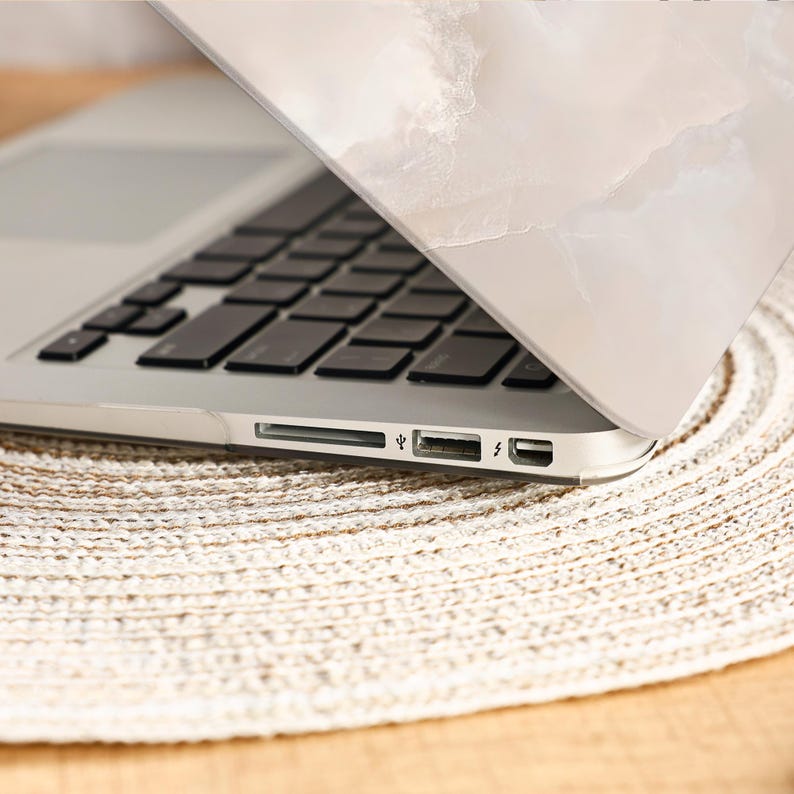 Peut inclure: Un ordinateur portable argent&eacute; avec un clavier noir, partiellement ouvert, r&eacute;v&eacute;lant l'&eacute;cran. L'ordinateur portable repose sur un dessous de plat beige tiss&eacute;. Le couvercle de l'ordinateur portable pr&eacute;sente un motif marbr&eacute; blanc et beige.