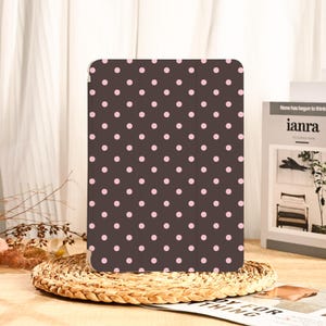 Pode incluir: Uma capa de tablet castanha com um padrão de bolinhas rosa. A capa é retangular e tem uma superfície lisa. O fundo inclui uma cesta de vime, um livro e uma cortina.