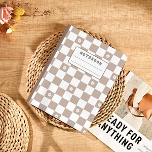 Puede incluir: Un cuaderno con un patrón de cuadros marrones y blancos y pequeños detalles florales. La palabra "NOTEBOOK" está impresa en la portada. El cuaderno descansa sobre un salvamanteles tejido, con una revista visible en el fondo.