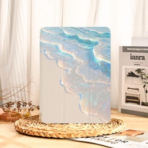 Può includere: Una custodia per tablet con un design ad acquerello di onde oceaniche nelle tonalità del blu, bianco e iridescente. La custodia è beige chiaro e ha una copertura magnetica. La custodia è su un sottobicchiere intrecciato.