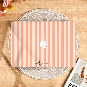 Peut inclure: Une coque d'ordinateur portable à rayures verticales pêche et blanches. Le logo Apple est visible au centre, avec le nom "Alison" en écriture cursive en dessous. La coque est posée sur un dessous de plat tressé.