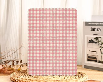 Può includere: Una custodia per tablet con un motivo a quadretti rosa e bianchi. La custodia è rettangolare e poggia su un sottobicchiere rotondo e intrecciato. Lo sfondo include una tenda bianca e un libro intitolato "ianra".
