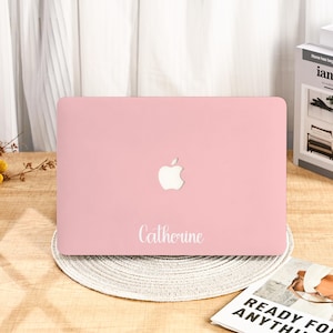 May include: A light pink laptop case with a white Apple logo and the name "Catherine" in a cursive font. The laptop case is resting on a round, woven placemat on a wooden surface.