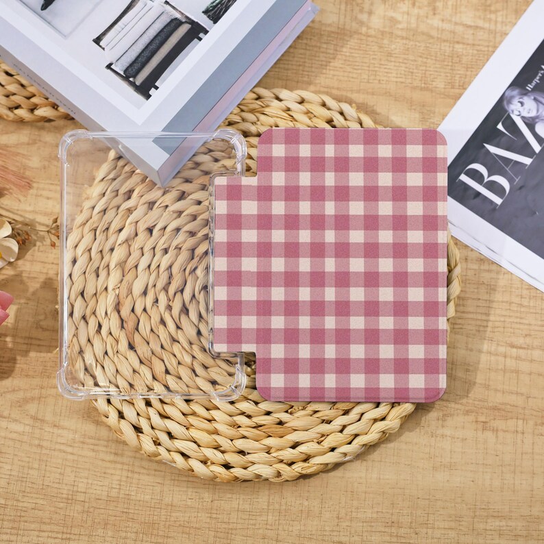 May include: A clear phone case and a pink and white checkered phone grip are displayed on a woven straw mat. A magazine and a book are partially visible in the background. The phone grip has a unique shape.