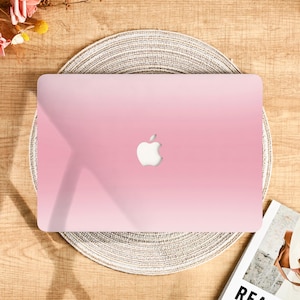 Può includere: Una custodia per laptop con sfumatura rosa e logo Apple bianco. La custodia è su un sottobicchiere intrecciato, con uno sfondo in legno. La custodia è in tinta unita e ha una finitura liscia.