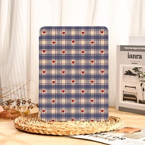 May include: A tablet case featuring a blue, beige, and grey plaid design, punctuated with small red hearts. The case is presented on a woven mat, with a book and other decorative items visible in the background.