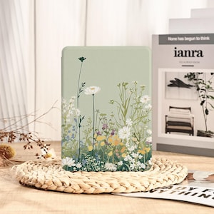 Può includere: Una custodia per tablet verde chiaro con un design floreale con fiori selvatici bianchi, gialli e viola. La custodia ha una forma rettangolare e poggia su un tappetino intrecciato. Il design include steli alti e petali delicati.