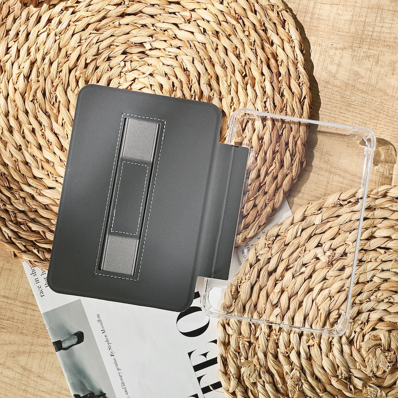May include: A gray tablet case with a built-in stand and a clear protective cover. The case is made of a smooth material and features a stitched design. The items are placed on a woven, circular mat.