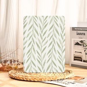 May include: A tablet case with a light green leaf pattern on a white background. The leaves are arranged in a repeating, diagonal pattern. The case is resting on a woven, circular placemat.