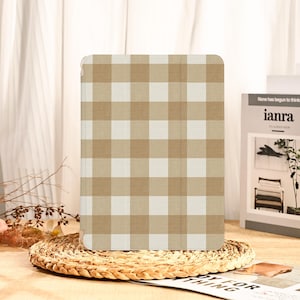 May include: A tan and white checkered tablet case. The case is rectangular with rounded corners and a grid pattern. The case is sitting on a woven placemat. A book with the text "ianra" is in the background.