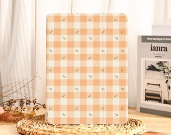 May include: A tablet case with a peach and white gingham pattern, adorned with small floral accents. The case is rectangular and has a folding cover. The case is resting on a woven placemat.
