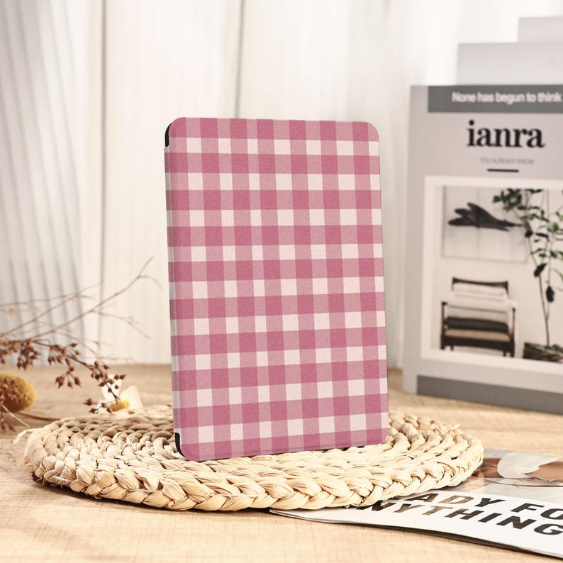 May include: A pink and white gingham pattern tablet case. The case is rectangular and has a black border. It is sitting on a woven placemat. In the background is a book with the text "ianra".