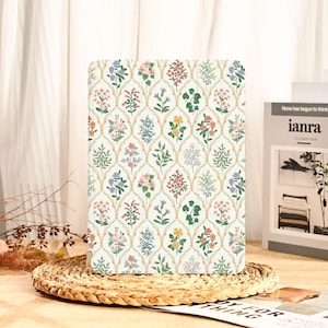 May include: A white tablet case with a floral pattern. The design features a repeating pattern of colorful flowers and green leaves within a gold-colored trellis design. The case is resting on a woven placemat.