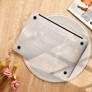 Peut inclure: Un ordinateur portable avec un &eacute;tui blanc &agrave; motif marbr&eacute;. L'&eacute;tui est translucide avec des pieds noirs et des fentes d'a&eacute;ration. L'ordinateur est pos&eacute; sur un dessous de plat rond et tress&eacute;, sur une surface en bois.