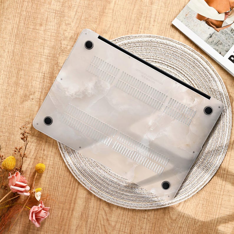 Peut inclure: Un ordinateur portable avec un &eacute;tui blanc &agrave; motif marbr&eacute;. L'&eacute;tui est translucide avec des pieds noirs et des fentes d'a&eacute;ration. L'ordinateur est pos&eacute; sur un dessous de plat rond et tress&eacute;, sur une surface en bois.