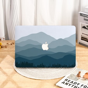 May include: A laptop case featuring a mountain range design in shades of blue and gray. The Apple logo is visible. The case is resting on a round, woven placemat. A magazine with the words "READY FOR ANYTHING" is visible.