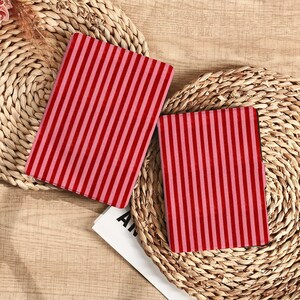 Può includere: Due quaderni rettangolari con strisce verticali rosse e rosa poggiano su sottopiatti intrecciati. I quaderni sono aperti, rivelando il motivo a righe. Lo sfondo è una superficie di legno marrone chiaro.