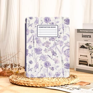 May include: A composition book with a floral design in shades of purple and white. The cover features a pattern of flowers and butterflies. The text "COMPOSITION BOOK" is printed on the front. The book is resting on a woven mat.