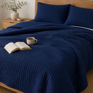 May include: A deep navy blue quilt featuring a subtle white dotted pattern and vertical stitched lines, paired with two matching rectangular pillow shams. An open book and a ceramic mug are placed on the surface of the textured bedding.