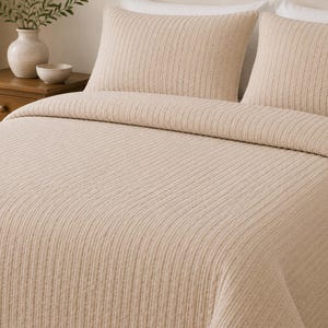 May include: A beige bedspread and two matching rectangular pillow shams featuring a vertical ribbed texture. The bedding set has a consistent light tan colour and a uniform stitched pattern that runs across the entire surface of the fabric.
