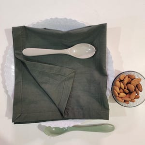 Op de afbeelding: Een donkergroene stoffen servet met een witte en een groene lepel op een wit oppervlak. Daarnaast een klein glazen schaaltje met amandelen. De compositie creëert een minimalistische esthetiek, met de lepels en het servet als focuspunten.