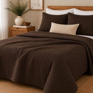 May include: A dark brown bedspread with a subtle vertical textured pattern, two matching dark brown pillow shams, and a single rectangular beige accent pillow. The bedding is arranged on a bed with white sheets and a wooden headboard.