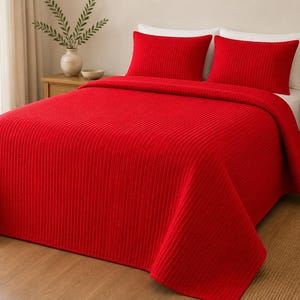 May include: A vibrant red bedspread featuring a vertical channel-stitched pattern that covers the entire surface. The set includes two matching rectangular red pillow shams placed against white pillows, creating a bold and uniform color display.