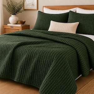 May include: A dark forest green quilt with a vertical stitched texture covering a bed. The bedding set includes two matching dark green pillow shams and a single rectangular cream-colored accent pillow placed in the center of the bed.