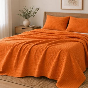 Op de afbeelding: Een feloranje sprei en twee bijpassende rechthoekige kussenslopen met een subtiel, getextureerd ruitpatroon. De beddengoedset is over een bedframe gedrapeerd en toont de heldere, effen kleur en de gewatteerde stofconstructie.