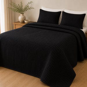 Op de afbeelding: Een effen zwarte sprei met een verticale ribbeltextuur die een bed bedekt. De set bevat twee bijpassende zwarte rechthoekige kussenslopen die tegen witte kussens zijn geplaatst, wat zorgt voor een uniforme donkere esthetiek voor het beddengoed.