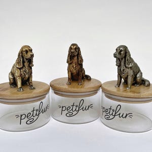Puede incluir: Tres tarros de cristal con tapas de bambú, cada uno coronado con una figura de perro. Los tarros tienen la palabra "petifur" en una fuente de escritura. Las figuras de perro son en tonos marrones y bronce.
