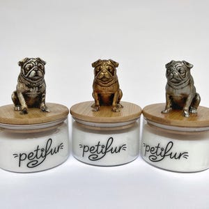 May include: Three glass jars with bamboo lids, each topped with a pug figurine. The jars are white with the word "petifur" in black script. The pug figurines are in bronze, silver, and gold tones.