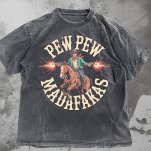 May include: Dark gray t-shirt featuring a cowboy on a horse, firing two guns with red bursts. The shirt has the text "PEW PEW MAUFAKAS" in a circular design. The shirt has a vintage, distressed look.