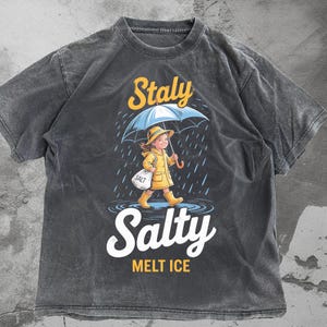 Pode incluir: Camiseta cinza escuro com um gráfico de uma pessoa de capa de chuva e botas amarelas segurando um guarda-chuva, com as palavras "Staly Salty Melt Ice". A pessoa está carregando uma sacola com a palavra "Salt".