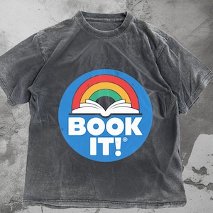 May include: A dark grey t-shirt with a circular graphic. The graphic features a rainbow arch over an open book, set on a blue background with the words "BOOK IT!" in white. The shirt has a washed, vintage appearance.